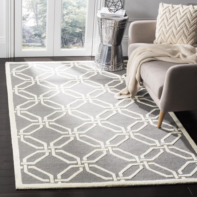 Safavieh Cambridge Kierra Geometric Area Rug featuring a grey wool base with an off-white geometric chain-link pattern, placed on dark hardwood floors in a living room with a beige sofa, knitted throw, and metallic side table, ideal for adding modern American style and texture to accent rug areas.