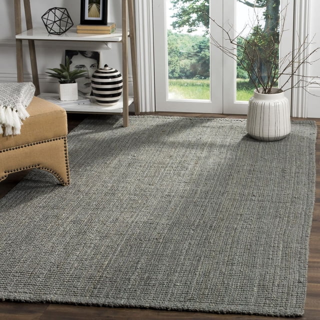 SAFAVIEH Natural Fiber Milica Braided Jute Area Rug in grey with a durable braided texture on a hardwood floor by glass doors overlooking greenery, styled with a beige nailhead-trimmed chair, a white shelf with decorative items including a striped vase, and a white ceramic pot holding bare branches, ideal for coastal or casual beach house décor.