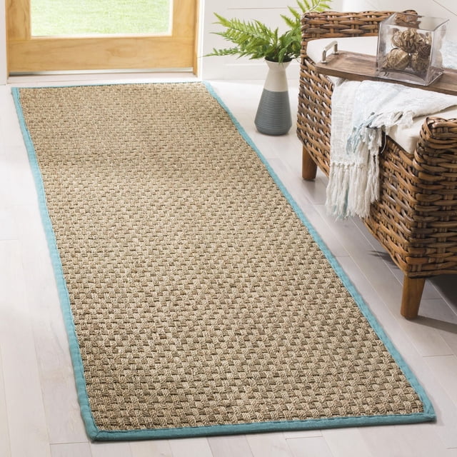 Safavieh Natural Fiber Arbor Border Area Rug with woven sisal texture and light blue fabric border, placed on light hardwood flooring beside a wicker chair with a white and blue throw and a green plant in a vase, ideal for coastal or casual beach house style and suitable as a hallway runner.