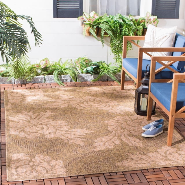 Safavieh Courtyard Adrian Floral Outdoor Area Rug in beige and light brown with a large floral pattern, made of durable polypropylene sisal weave for outdoor patios. It is placed on a wooden deck with two wooden chairs featuring blue cushions and a white starfish pillow, a black lantern, and blue canvas shoes nearby. The setting includes green plants and a white wall with black shutters. The rug is also suitable for high-traffic indoor areas.