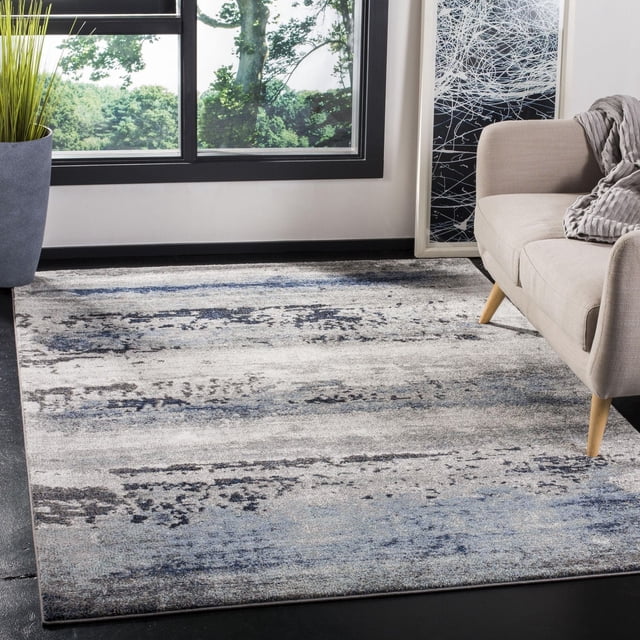Safavieh Galaxy Tapis Abstrait Jacques Mettant en Vedette un Motif Abstrait d'Inspiration Céleste dans des Tons Bleu, Gris et Noir avec une Poil Doux. Exposé dans un Salon Contemporain sur un Plancher Sombre à Côté d'Un sofa beige avec des Pieds en Bois, Près d'Une Grande Fenêtre Surplombant des Arbres Verts. Idéal pour Accentuer la Déco Maison Moderne.