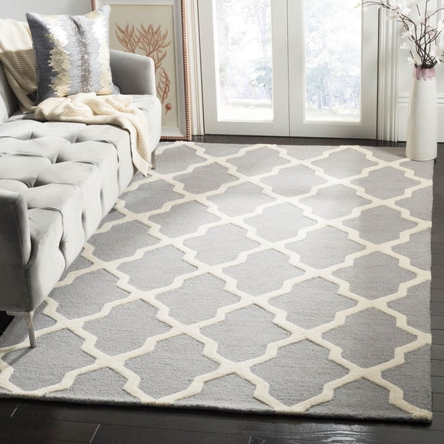 Safavieh Cambridge Liam Geometric Area Rug featuring hand-tufted wool with a grey base and cream geometric lattice pattern, placed on dark hardwood flooring in a living room with a grey tufted sofa, cream throw, and decorative pillow near glass doors and a white vase with branches, ideal for adding texture and modern American style to home decor.