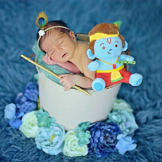 MODI TOYS Krishna (Small - 6 inch) Mantras Singing Plush displayed in a decorative setting. The plush features a blue Baby Krishna figure with a flute in his left hand, a blessing gesture with his right hand, and a peacock feather on top. It is surrounded by a floral arrangement and placed in a light-colored container. The toy is designed to sing five Krishna mantras and includes a velcro strap for easy attachment to strollers, diaper bags, or other items.