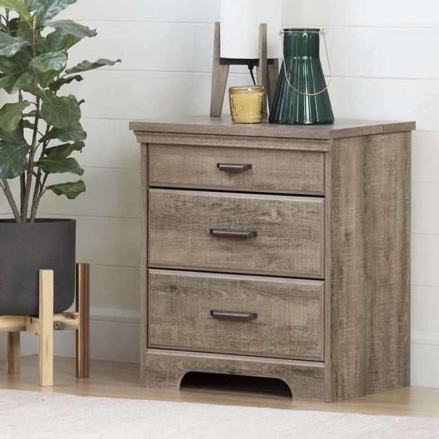 South Shore Versa Nightstand with Charging Station and Drawers for Adult in weathered oak finish, featuring three drawers with metal handles and a sliding top shelf that hides a power bar for device charging, placed on a light wood floor next to a green leafy plant in a black planter with wooden stand, against a white shiplap wall, ideal for bedside storage and discreet electronics charging.
