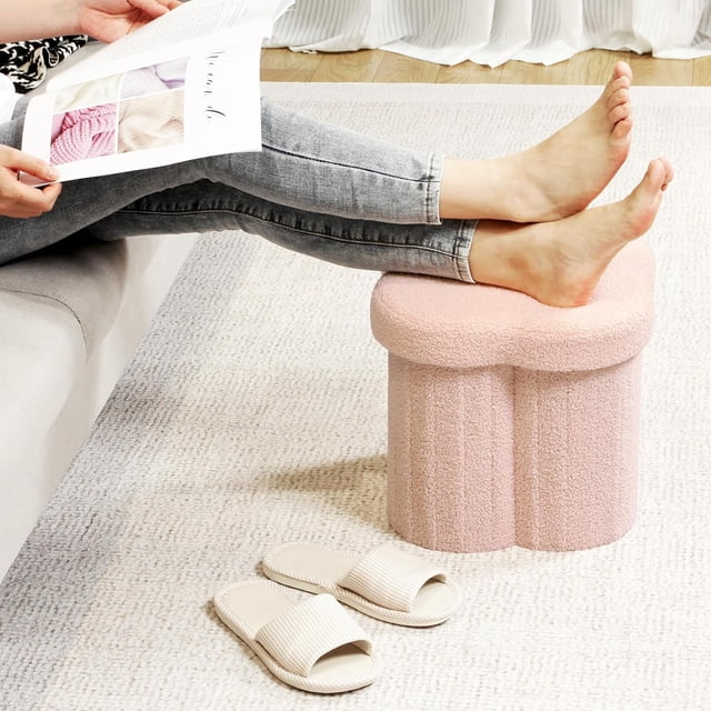 PINPLUS 12.6" pink ottoman storage box in a flower shape, crafted from teddy velvet fabric, displayed in a cozy living room. The foldable design functions as a footstool, toy chest, or extra seating. A person rests their feet on the ottoman, showcasing its versatility.