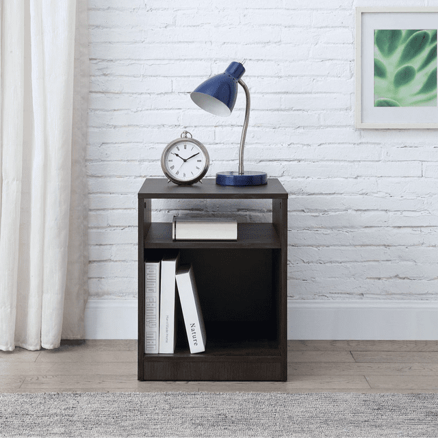 Mainstays table de Chevet à Étagères Ouvertes en Fini Chêne Foncé, une table de Chevet Compacte avec une surface Supérieure Tenant une Lampe de Bureau Bleue et un Réveil Argenté, une Étagère Centrale avec un Livre Fermé, et un Grand Casier Inférieur Rangeant Trois Livres Verticaux, Placée sur un Plancher de Bois Clair contre un Mur de Briques Blanches avec des Oeuvres d'Art Abstraites Encadrées Vertes, Idéale pour les Chambres d'Enfants et le Rangement de Chevet.