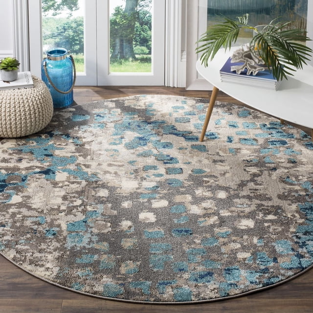 Safavieh Monaco Driskoll Abstract Area Rug, a durable round accent rug with blue, grey, and beige abstract patterns in a power-loomed polypropylene weave featuring high and low loop pile, placed on a wooden floor near glass doors with modern decor including a white table and green plant. Suitable for adding Bohemian-chic flair to living spaces.