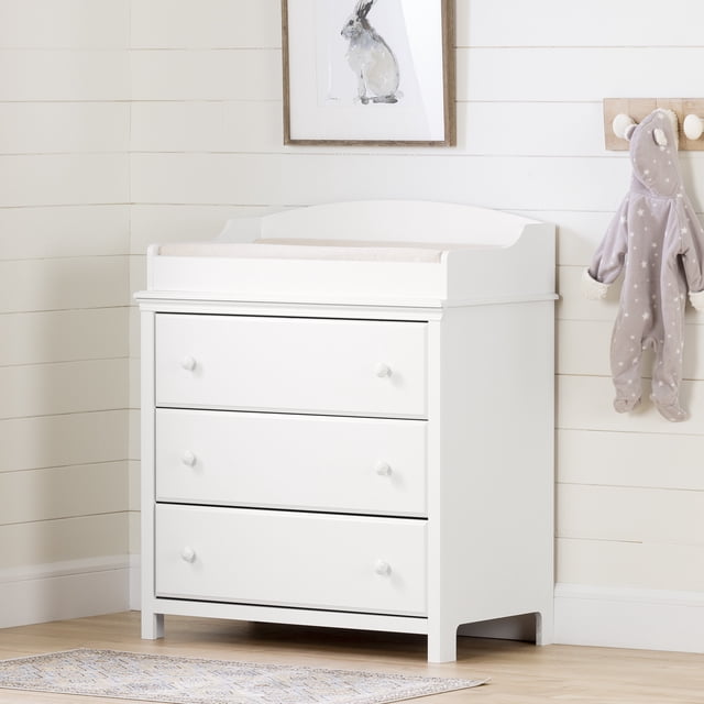 South Shore Cotton Candy 3-Drawer Changing Table for Infant in pure white with three large drawers and white ceramic knobs, compact design with beveled top edge and raised sides for changing pad, ideal for small nursery spaces, shown against white shiplap walls with framed rabbit art and hanging baby outfit on light wood floor.