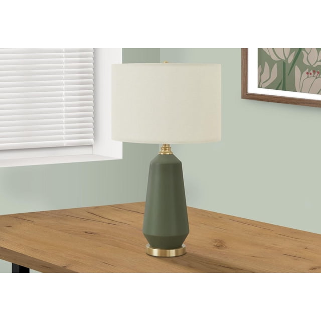 Monarch Specialties Lighting 26"h table lamp with a green ceramic tapered base and gold metal accents, featuring an ivory linen drum shade, placed on a wooden side table near a window with white blinds and a framed botanical print on a pale green wall, ideal for ambient lighting in living rooms or bedrooms.