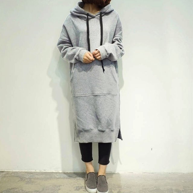 Nouvelle Robe Longue Ample pour Femmes à Capuche en Gris, avec Capuche à Cordon, Poche Kangourou, Fente Latérale et Manches Longues avec Épaules Tombantes. Fabriquée en Tissu Doux en polyester et spandex, Offre Chaleur et Confort. la Robe a des Poignets et un Ourlet Côtelés, un Motif Uni, et un style Décontracté et Tendance. Montrée dans une Vue Frontale, Assortie à des leggings Noirs et des Chaussures Grises à Enfiler.