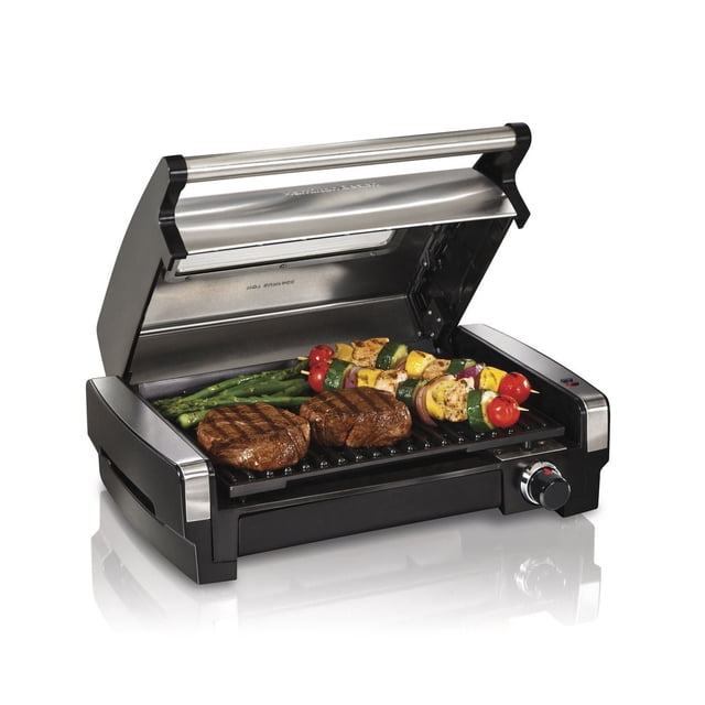 Hamilton Beach Indoor Electric Searing Grill with Viewing Window, stainless steel housing and black base, open lid revealing two seared steaks, vegetable skewers with cherry tomatoes, zucchini, onions, and asparagus on the nonstick grill plate, featuring an adjustable temperature control knob on the front right side.