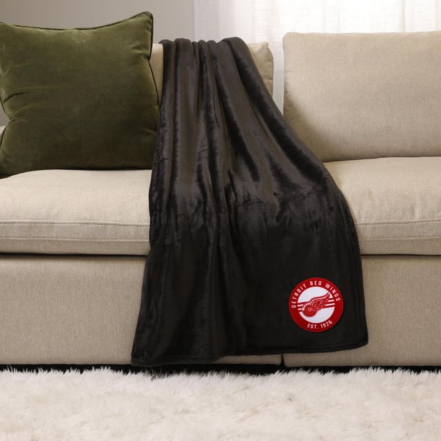 NHL, NBA, MLB Black Sports Throw Blanket, 50" x 60", draped over a beige sofa with a green cushion, made of soft plush 100% polyester fabric featuring a red Detroit Red Wings logo patch, ideal for indoor use and cozy warmth.
