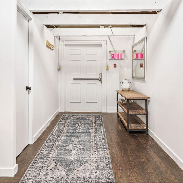 ECARPET Vintage Oriental Area Rug for Living Room Bedroom, Oregon Bijar Collection in a white hallway with exposed ceiling beams and dark hardwood floors. The rug features a traditional vintage pattern in grey and beige tones, centered on the floor. A rustic wooden console table with metal frame and white ceramic lamp sits beneath a rectangular mirror on the right wall. A white door with decorative trim and a pink 'SORTIE' exit sign is at the hallway's end. The rug is also suitable for accentuating narrow entryways or corridors.