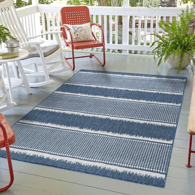 Erbanica Quick Dry Indoor/Outdoor Rug, 8 ft x 11 ft, displayed on a patio with a modern blue and white striped design. The rug features a water and mildew-resistant surface, UV protection, and a durable, fade-resistant polypropylene construction. Suitable for high-traffic areas, patios, and decks, it is easy to clean by wiping with a damp cloth or hosing down. The setting includes outdoor furniture and plants, emphasizing its use in outdoor spaces.