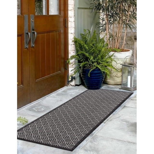 ECARPET Eve Contemporary Runner Rug with a geometric basketweave pattern in charcoal and light grey polypropylene fibers, placed on a stone floor outside wooden double doors, accompanied by potted green plants and a metal lantern, suitable for indoor and outdoor use and ideal for entryways.