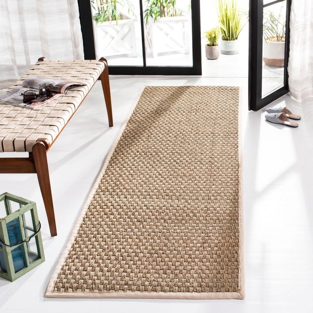 Safavieh Natural Fiber Arbor Border Area Rug with woven sisal texture and fabric border in natural beige, placed on a white floor near a woven bench and open glass door with potted plants, ideal for coastal or casual beach house style and durable hallway runner use.