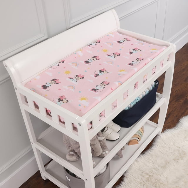 Disney Change Pad Cover in pink with Minnie Mouse, rainbows, suns, and clouds, fitted with elastic on a white wooden changing table with two shelves holding baby shoes, plush toys, and storage bins in a nursery with white walls and hardwood floor.