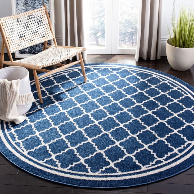 SAFAVIEH Amherst Trina Geometric Area Rug, round with a blue and white geometric lattice pattern, made of durable polypropylene for indoor-outdoor use, placed on dark hardwood floor with a woven wooden chair, a white basket holding a beige throw, and a green potted plant near a bright window with beige curtains.