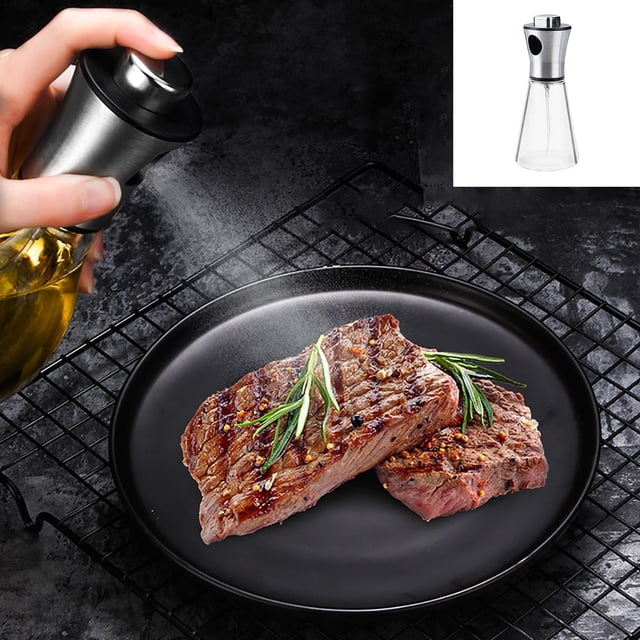 WIFKLSIIPG 200ml glass and stainless steel cooking oil spray bottle, shown in use spraying oil onto grilled steak garnished with rosemary on a black plate. The bottle features a clear glass body and a stainless steel top with a pressure nozzle for controlled oil dispensing. Suitable for olive oil, rapeseed oil, vinegar, and other liquids, ideal for air fryers, barbecues, and general cooking.