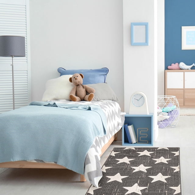ECARPET Kids Room, Nursery, Playroom, Bedroom Area Rug, Angel Collection in grey with white star pattern, placed beside a child's bed with blue knit blanket, neutral pillows, and a teddy bear in a bright kids' room with blue and white decor, suitable for high traffic and pet-friendly areas.