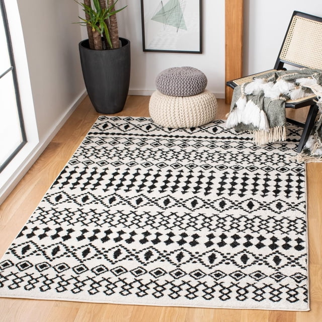 SAFAVIEH Tulum Herbert Distressed Southwestern Area Rug with black geometric and diamond patterns on a cream background, flat-woven synthetic yarn construction, placed on light hardwood flooring near a black planter, two knitted poufs, and a cane chair with a fringed throw, suitable for enhancing rustic and boho living spaces.