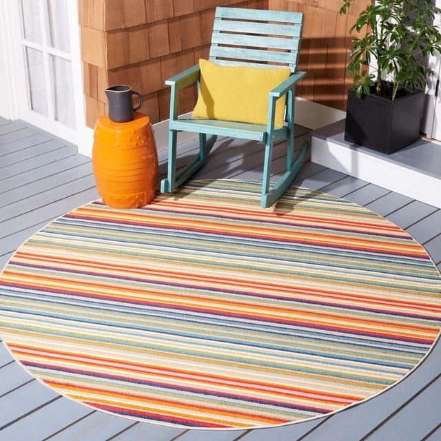 Safavieh Cabana Siomha Striped Outdoor Area Rug, round with multicolored horizontal stripes in red, blue, yellow, and green, placed on a light gray wooden patio deck. A light blue wooden rocking chair with a yellow cushion sits on the rug beside an orange ceramic side table holding a black pitcher. The rug is weather-resistant and ideal for outdoor patio use.
