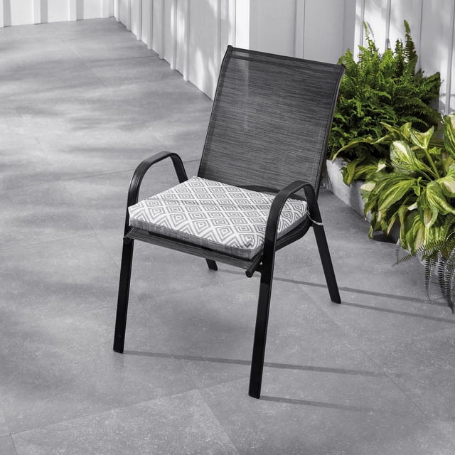 Mainstays Ensemble de 2 Coussins de Siège pour Chaise d'Extérieur en Métal Noir avec Dossier en Maille, Comprenant un Coussin Gris avec Motif Géométrique Blanc en Losanges, Placé sur un patio Carrelé Gris à Côté de Plantes Vertes et d'Une Clôture Blanche, Fabriqué avec un Tissu Hydrofuge Traité aux UV Idéal pour le Confort d'Assise Extérieur.