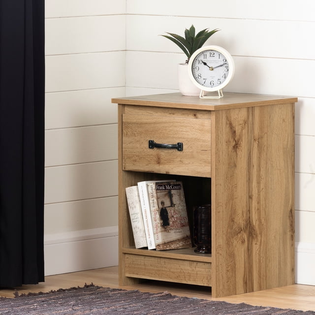 South Shore Tassio 1-Drawer Nightstand with Open Storage Space for Adult in Nordik Oak, front view showing a single drawer with antique style metal handle, open shelf holding books and a dark glass, topped with a white round clock and a potted plant, placed against white shiplap walls and light wood flooring, often used for bedside storage and display.