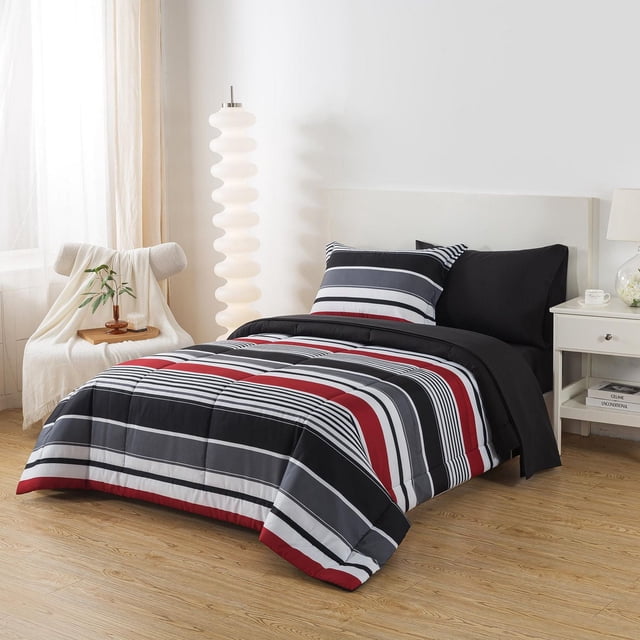 Mainstays Bed-in-a-Bag twin set with a striped comforter in black, white, gray, and red horizontal stripes, including matching striped and solid black pillowcases, displayed in a modern bedroom with light wood flooring, white headboard, white nightstand with lamp and books, and a cream throw-covered chair beside a white sculptural floor lamp, suitable for year-round comfort and easy bedroom coordination.