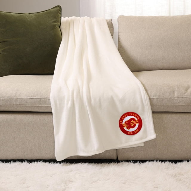 NHL, NBA, MLB White Sports Throw Blanket, 50" x 60", draped over a beige sofa with a green cushion, made of soft plush 100% polyester featuring a red Calgary Flames logo patch, ideal for indoor warmth and suitable for outdoor use on cool days.
