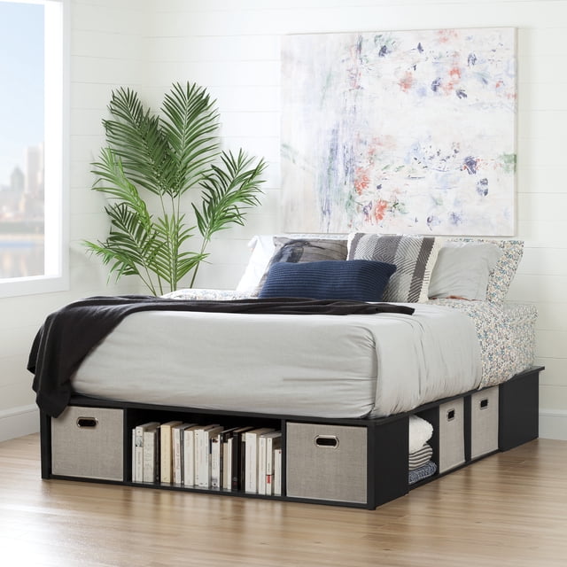 South Shore Flexible King Storage Platform Bed with Baskets in Black Oak, featuring a sturdy black oak frame with underbed storage compartments including fabric bins and open shelves holding books and linens. The bed is dressed in light grey sheets, floral pillowcases, and decorative pillows in navy and grey. Set in a bright room with white shiplap walls, light wood flooring, a large abstract painting above the headboard, and a tall green plant near a window with a city view. This platform bed requires no box spring and is suitable for maximizing storage in small bedrooms.