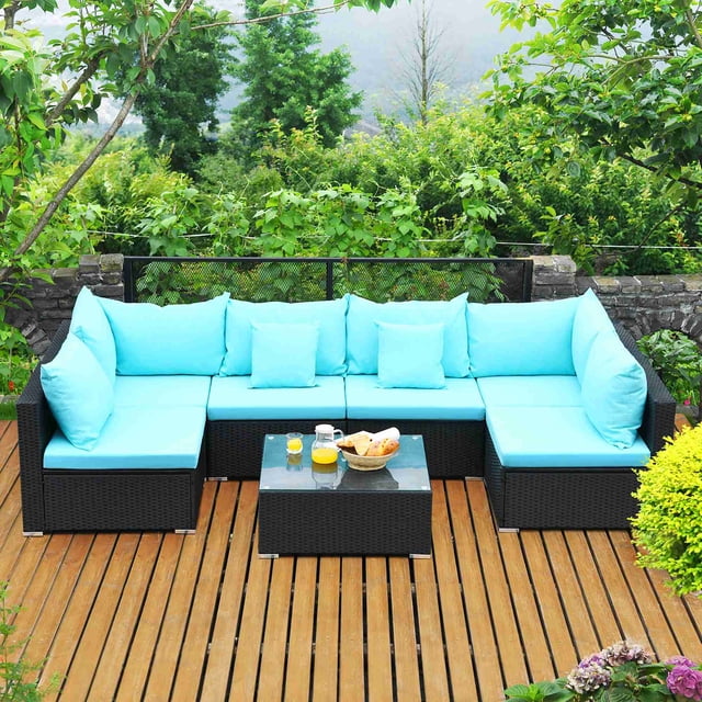 Costway 7PCS Patio Wicker Sofa Set Sectional Conversation Furniture Set in navy blue, ideal for outdoor spaces like gardens, patios, or poolside. Includes four armless sofas, two corner sofas, and a coffee table with a tempered glass top. Features a durable PE wicker frame, thick cushions, and pillows for comfort, arranged on a wooden deck surrounded by lush greenery.
