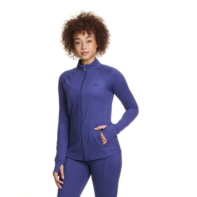 Reebok Women’s Reflex Performance Full-Zip Jacket with Pockets in purple, front view on a model wearing matching leggings. The jacket features a high collar, full zipper, thumbhole cuffs, and zippered invisible side pockets, made from stretchy moisture-wicking Motion Poly/Span Interlock fabric, ideal for workouts or layering.