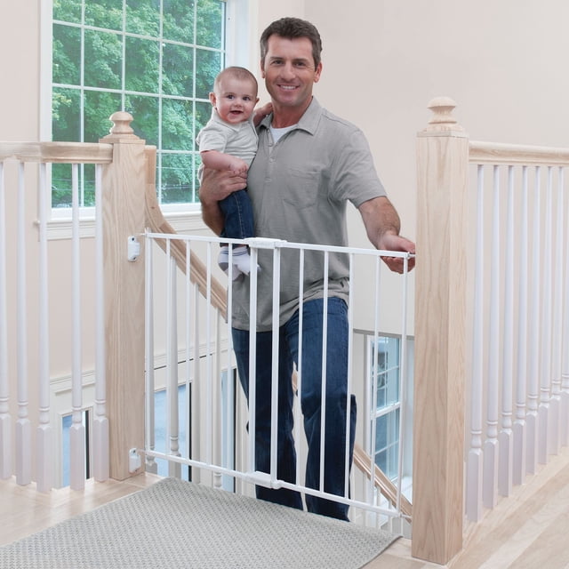 Safety 1st Top of Stairs Expanding Metal Gate installed at the top of a light wood staircase with white spindles, showing a white metal gate with vertical bars and a one-hand release lever being opened by an adult holding a toddler, designed to secure child-safe areas and prevent stair access.