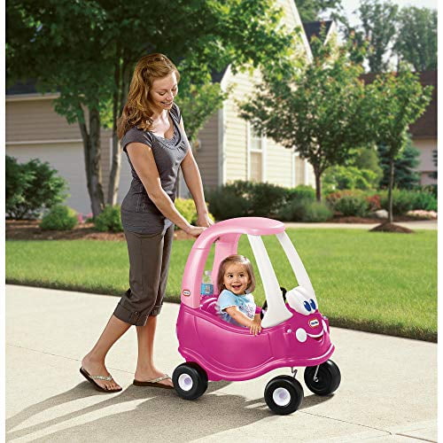 Little Tikes Princess Cozy Coupe Ride-On Toy in pink, featuring a toddler-sized car with working doors, a steering wheel, a horn, a gas cap, and an ignition switch. The toy is designed for boys and girls, shown being pushed by an adult on a sunny sidewalk in a residential neighborhood.