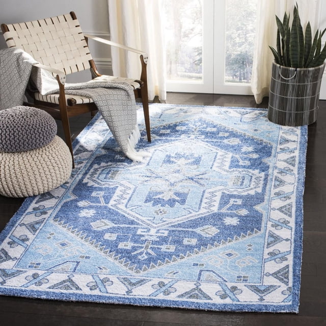 Safavieh Saffron Magdalena Tapis Oriental à Bordure en Coton Bleu et Blanc avec Motif floral Géométrique, Posé sur un Plancher de Bois Franc Sombre dans une Pièce Lumineuse Comprenant une Chaise à Sangles Tressées, des poufs Tricotés et une Plante Serpent en Pot, Convient comme Tapis d'Accent pour les Espaces de Vie.