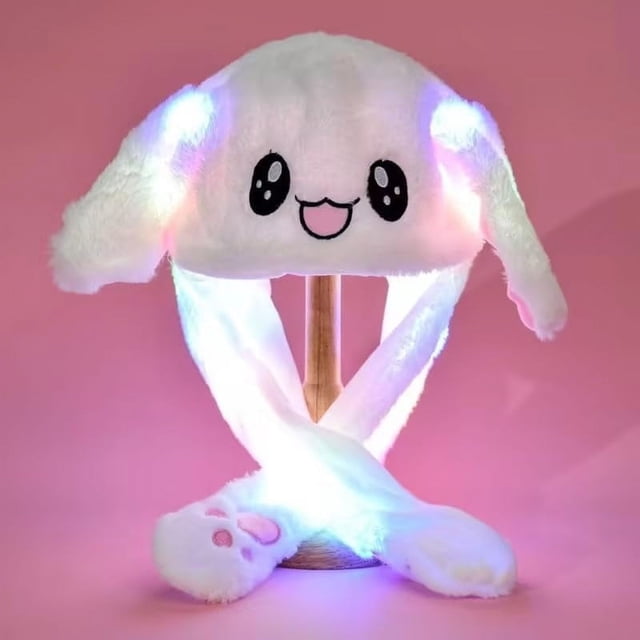 Luminous Bunny Hat with Ears, a cute plush toy warm hat featuring a rabbit design with moving ears. The hat is white with a smiling face and glowing LED lights in the ears and straps. The interactive design allows the ears to move when the paw-shaped ends are pressed. Displayed on a wooden stand against a pink background.