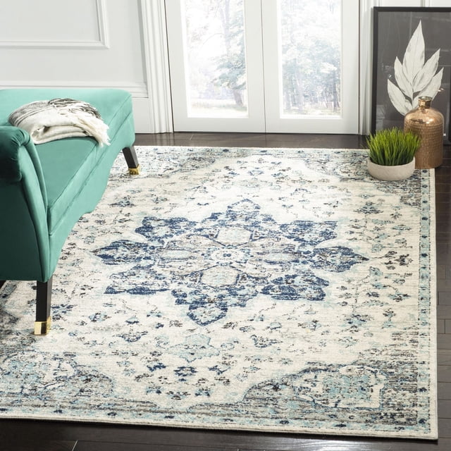 Safavieh Madison Louise Overdyed Floral Area Rug featuring a central blue floral medallion on a cream background, power-loomed with durable yarns for long-lasting use, displayed on dark hardwood floor beside a teal bench with gold-tipped legs near glass doors and decorative plants, suitable for transitional home decor.