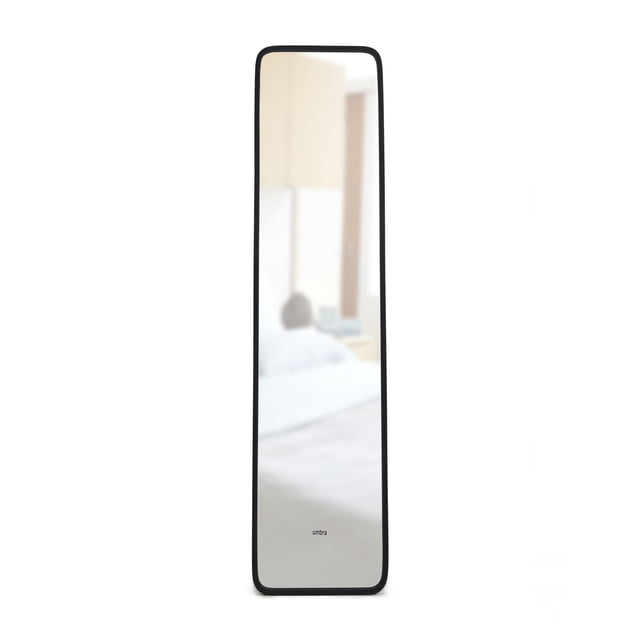 Umbra Hub Miroir Mural Pleine Longueur Noir avec Cadre Mince en Caoutchouc, Mesurant 62 par 14,5 Pouces, Montré Orienté vers l'Avant avec un Reflet Flou dans la Chambre, Conçu pour Être Fixé au Mur Ou Appuyé pour Égayer et Agrandir les Espaces. Sa Conception Légère le Rend Aussi Adapté aux Petits Espaces Ou aux Espaces Maladroits.