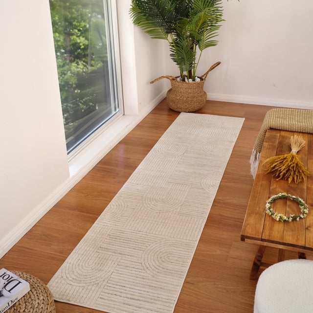 Rug Branch Astra Modern Indoor Area Rug in cream beige with geometric abstract patterns, shown as a runner on a hardwood floor beside a large window. Made of polyester with jute backing, it features linear and curved designs. Positioned near a woven basket planter with green foliage and a wooden bench with a yellow throw and dried flowers, suitable for hallways, kitchens, or living rooms.
