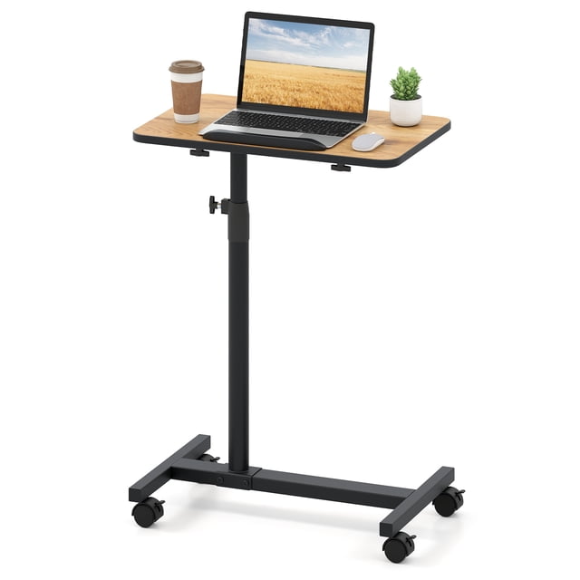 Giantex Adjustable Tilt Overbed Table in a natural wood finish with a black H-shaped steel base. The table features a 20.5" x 13" tiltable and 360° swivel tabletop, adjustable height from 27" to 44", and four smooth-rolling wheels, two of which are lockable. The table is shown with a laptop, coffee cup, and a small plant, demonstrating its use as a bedside or laptop desk. Rounded corners and an anti-slip edge ensure safety and stability.