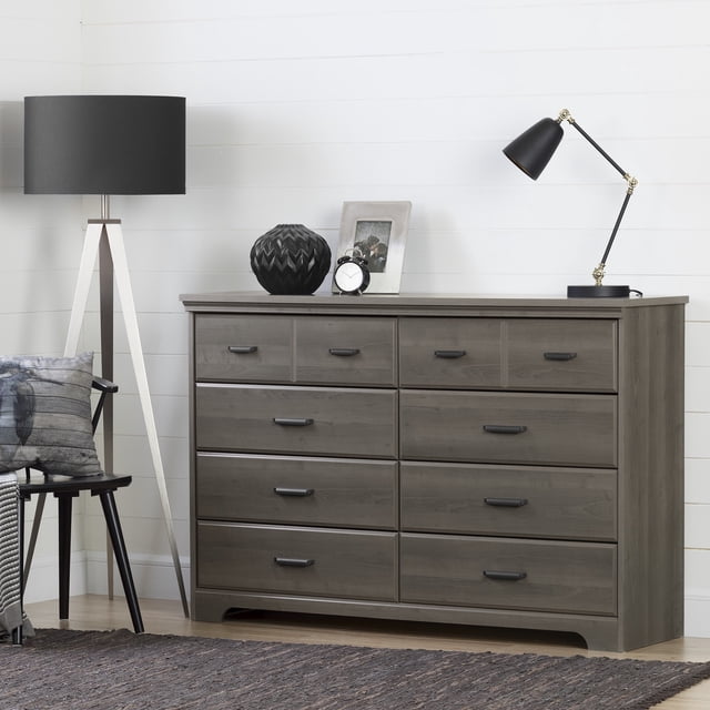 South Shore Versa 8-Drawer Dresser for Adult in gray maple finish with eight spacious drawers featuring black metal handles, shown front-facing in a bright room with white shiplap walls. The dresser top holds a black geometric vase, a small round clock, a photo frame, and a black adjustable desk lamp. Positioned beside a tripod floor lamp and a black chair with a gray cushion on a dark textured rug, this dresser offers practical storage and classic style, also suitable for organizing office supplies or craft materials.