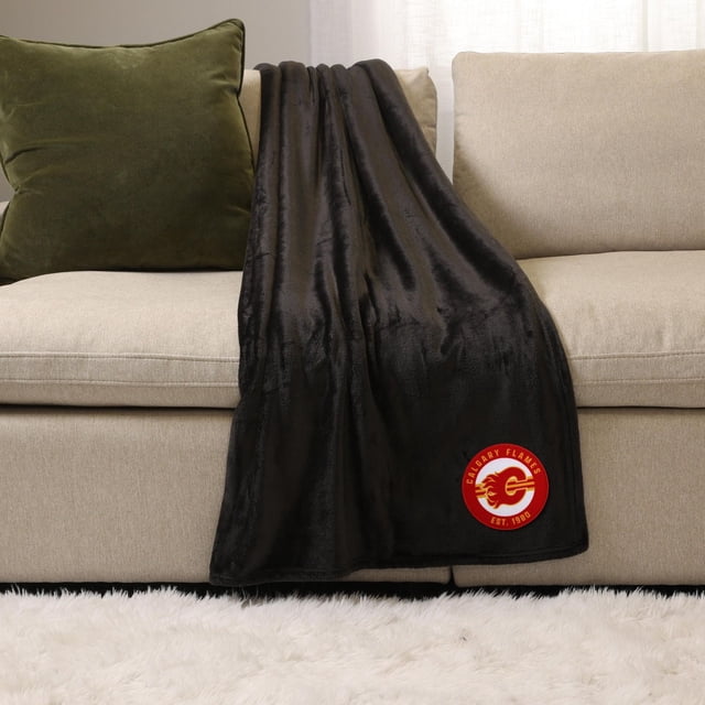 NHL, NBA, MLB Black Sports Throw Blanket, 50" x 60", plush 100% polyester blanket draped over a beige sofa with a green cushion, featuring a red Calgary Flames logo patch on the lower corner, suitable for indoor warmth and lounging.