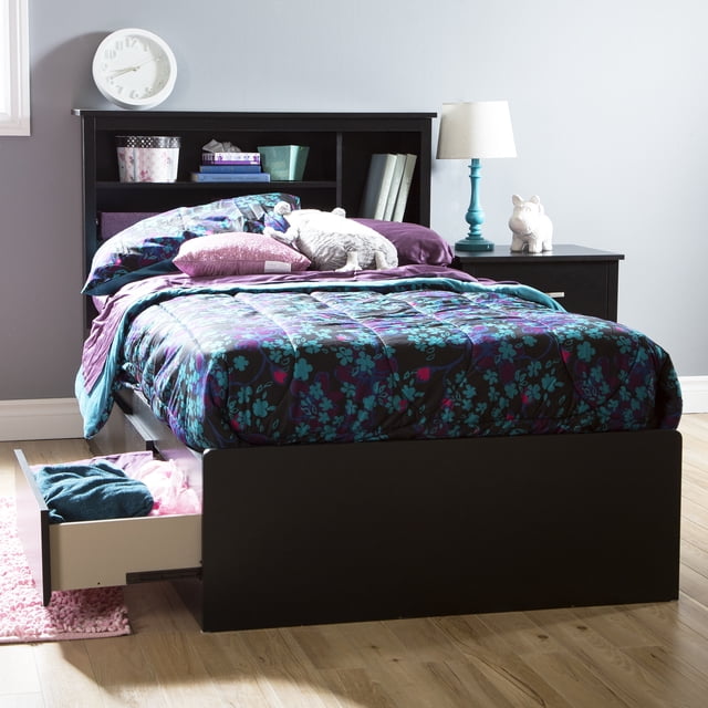 South Shore Step One Twin Mates Bed with 3 Drawers in Pure Black, shown in a front 3/4 view with one drawer open displaying folded clothes. The platform bed features a built-in headboard with shelves holding books and storage boxes. It is dressed with a floral comforter in purple, teal, and blue tones, with pillows and stuffed toys on top. A black nightstand with a turquoise lamp and white decorative figure sits beside the bed on a light wood floor against a grey wall. The drawers have metal slides and can be positioned on either side for flexible storage.