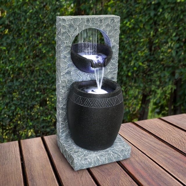 Angelo Décor Cascata Fountain, a 32-inch tall outdoor water fountain made from durable Comp-Cast stone, fibre, and resin. It has a textured grey frame with a circular cutout where water flows from a smaller black bowl with geometric patterns into a larger black basin below. Includes energy-efficient pump and LED lighting, suitable for patio decor and creating a calming ambiance.