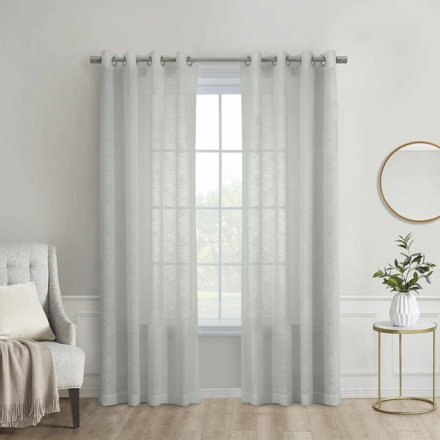 Loft Living Banan Sheer Grommet Single Curtain Panel 52 x 108 in Light Grey made of 100% polyester sheer fabric hanging on a metal rod, softly filtering natural light while providing daytime privacy, displayed in a minimalist room with a beige armchair and a round side table holding a white vase with green foliage, suitable for modern window coverings and light-diffusing treatments.