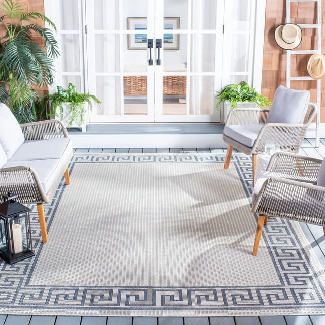 SAFAVIEH Bermuda Isidora Geometric Machine Washable Indoor/Outdoor Area Rug on a white wooden deck patio. The beige rug features a dark blue Greek key border and textured weave. It is surrounded by light grey cushioned outdoor chairs, green potted plants, a black candle lantern, and a wooden wall with straw hats. This durable, weather-resistant rug is ideal for patios, decks, and porches, often used to enhance outdoor living spaces.