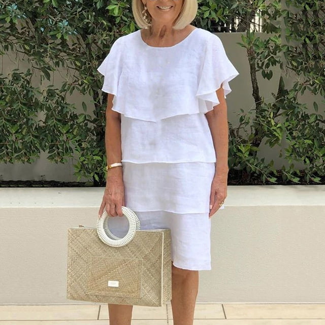 Gspmoly Robes pour Femmes Grande Taille, une Robe Décontractée Unie à Col Rond Montrée en Blanc. la Robe Présente des Manches Courtes avec des Volants Superposés et un Ourlet Longueur Genou. Elle Est Conçue pour un Décor Estival, Assortie à un Sac à Main Tissé et à des Accessoires Dorés, avec un Fond Extérieur Vert Luxuriant.