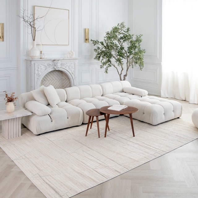 Rug Branch Astra Modern Indoor Area Rug in beige cream with abstract geometric pattern, displayed in a bright living room featuring a tufted cream sectional sofa, two round wooden nesting tables, a white decorative fireplace with minimalist art, and a tall green indoor plant near sheer white curtains.