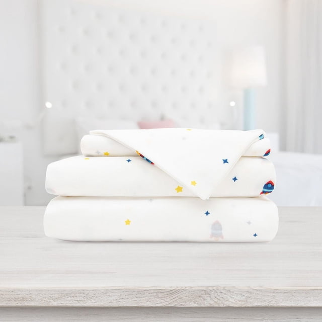 Safdie & Co. Premium ultra soft printed sheet set folded, featuring stars and rockets on white fabric, displayed on a light wood table in a bright bedroom.